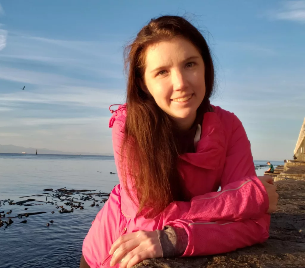 Nicole, in a pink sweater, leans forward on a rock with a rocky beach and water behind her.